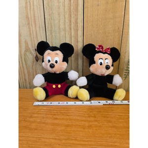 K&ouml;nnte beinhalten: Zwei Pl&uuml;schpuppen von Mickey Mouse und Minnie Mouse. Mickey tr&auml;gt rote Shorts, ein schwarzes Hemd und gelbe Schuhe. Minnie tr&auml;gt eine rote Schleife, ein schwarzes Kleid und gelbe Schuhe. Beide haben schwarze Ohren und wei&szlig;e Handschuhe.