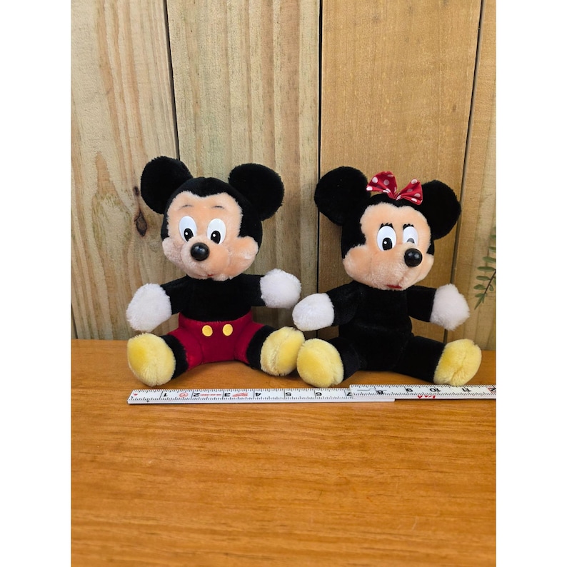 K&ouml;nnte beinhalten: Zwei Pl&uuml;schpuppen von Mickey Mouse und Minnie Mouse. Mickey tr&auml;gt rote Shorts, ein schwarzes Hemd und gelbe Schuhe. Minnie tr&auml;gt eine rote Schleife, ein schwarzes Kleid und gelbe Schuhe. Beide haben schwarze Ohren und wei&szlig;e Handschuhe.