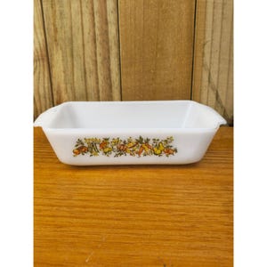 May include: White rectangular casserole dish with handles, decorated with a colorful harvest-themed pattern of vegetables and fruits. The dish is made of glass and sits on a wooden surface.
