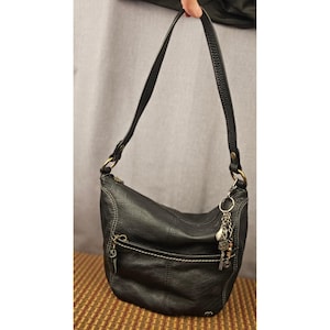 May include: A black hobo-style shoulder bag with a textured surface, featuring a single strap attached with brass-toned rings, a front zippered pocket with contrast stitching, and a decorative metal charm hanging from the side hardware.