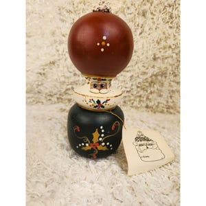 May include: A three-tiered Christmas ornament featuring a dark red sphere, a Santa Claus face, and a black base with gold and red accents. A tag with a Santa illustration hangs from the ornament.