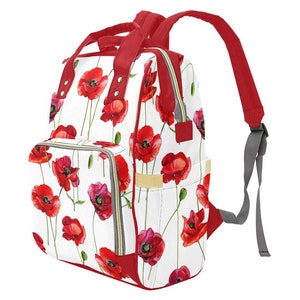 Puede incluir: Mochila de pañales con estampado floral de amapolas rojas sobre fondo blanco. La mochila tiene una cremallera roja y una correa ajustable gris.
