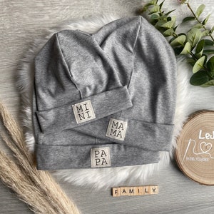 Könnte beinhalten: Drei graue Strickmützen mit den Worten "MI NI", "PA PA" und "MA MA", die darauf gestickt sind. Die Mützen liegen auf einer weißen, flauschigen Oberfläche mit einem Holzschild, auf dem "LeJaMo Handmade" steht.