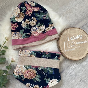 Könnte beinhalten: Ein florales Set aus Mütze und Halstuch in den Farben Rosa, Beige und Blau. Die Mütze hat einen rosa umgeschlagenen Rand und ein Schleifendetail. Der Halstuch hat einen beigefarbenen Streifen.