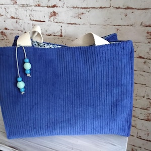 Könnte beinhalten: Eine blaue Cord-Tragetasche mit cremefarbenen Henkeln. Die Tasche hat eine vertikale Rippenstruktur und ist mit einer Kette aus blauen und türkisfarbenen Perlen verziert. Die Tasche steht auf einer weißen Oberfläche.
