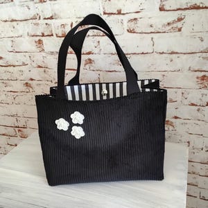 Könnte beinhalten: Schwarze Cord-Tragetasche mit schwarzen Henkeln und einem weiß-schwarz gestreiften Innenraum. Die Tasche hat drei weiße Blumenapplikationen auf der Vorderseite. Die Tasche steht auf einer weißen Oberfläche vor einer Backsteinmauer.