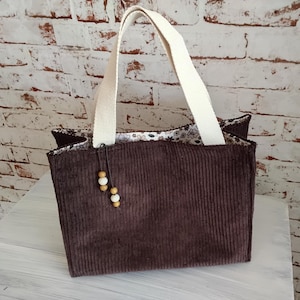 Könnte beinhalten: Eine braune Cord-Tragetasche mit cremefarbenen Henkeln. Die Tasche hat ein floral gemustertes Futter und einen dekorativen Holzperlen-Anhänger. Die Tasche hat eine rechteckige Form und eignet sich zum Tragen verschiedener Gegenstände.