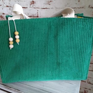 Könnte beinhalten: Eine grüne Cord-Tragetasche mit weißen Canvas-Griffen. Die Tasche hat eine rechteckige Form und eine vertikale Rippenstruktur. Dekorative Holzperlen hängen von oben. Die Tasche steht auf einer weißen Holzoberfläche.