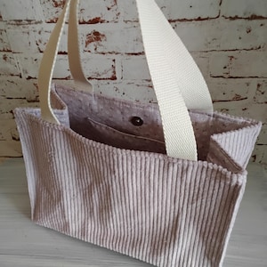 Könnte beinhalten: Eine hellbeige Cord-Tragetasche mit cremefarbenem Riemen. Die Tasche hat eine rechteckige Form mit vertikalen Cordlinien. Das Innenfutter ist ein hellrosa Stoff mit kleinem Blumenmuster. Ein Knopfverschluss ist sichtbar.