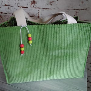 Könnte beinhalten: Eine grüne Cord-Tragetasche mit cremefarbenen Henkeln. Die Tasche hat eine dekorative Schnur mit roten, gelben und grünen Perlen. Die Tasche steht auf einer weißen Oberfläche vor einer Backsteinmauer.