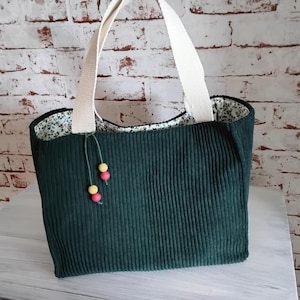 Könnte beinhalten: Dunkelgrüne Cord-Tragetasche mit cremefarbenen Henkeln. Das Innere der Tasche hat ein Blumenmuster. Gelbe und rosa Holzperlen hängen an der Vorderseite der Tasche.