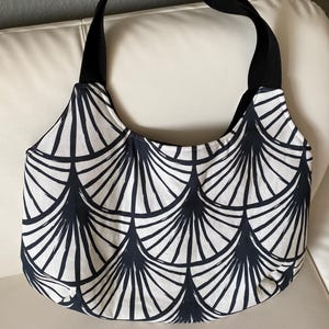 Könnte beinhalten: Eine Handtasche mit weißem und marineblauem Muster und schwarzen Trägern. Die Tasche hat ein sich wiederholendes fächerartiges Design. Die Träger bestehen aus einem robusten schwarzen Material. Die Tasche liegt auf einer weißen Oberfläche.