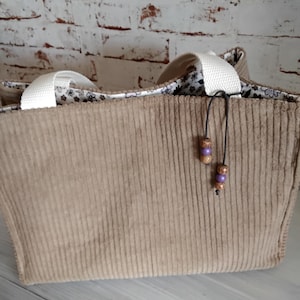 Könnte beinhalten: Eine beigefarbene Cord-Tragetasche mit weißen Henkeln. Die Tasche hat ein Futter mit Blumenmuster und eine dekorative Schnur mit lila und braunen Perlen. Die Tasche ist rechteckig und hat eine vertikale Rippenstruktur.