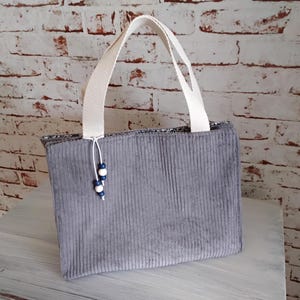 Könnte beinhalten: Eine rechteckige Handtasche aus grauem Cord mit einer texturierten vertikalen Rippenstruktur. Die Tasche verfügt über zwei stabile Tragegriffe aus weißem Webstoff und einen dekorativen Anhänger mit zwei kleinen weißen und zwei blauen Holzperlen.