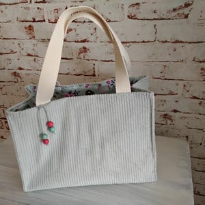 Könnte beinhalten: Hellblaue Cord-Tragetasche mit cremefarbenen Griffen. Die Tasche hat ein floral gemustertes Futter und ist mit roten und grünen Perlen verziert. Die Tasche steht auf einer weißen Oberfläche vor einer Backsteinmauer.