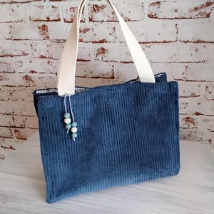 Könnte beinhalten: Eine rechteckige Tragetasche aus blauem Cord mit gerippter Textur, zwei stabilen, cremeweißen gewebten Tragegriffen und einem dekorativen Anhänger aus drei kleinen, runden blauen und weißen Perlen, der an der Basis eines Griffs befestigt ist.