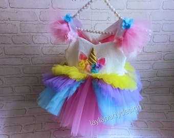 May include: A white unicorn tutu dress with rainbow tulle layers, pink and blue tulle sleeves, and a gold horn. The dress has a white bodice with a unicorn applique and is decorated with colorful flowers.
