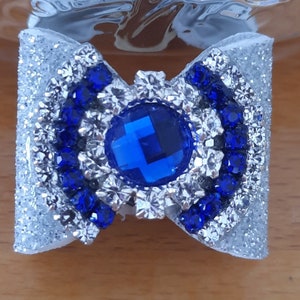 Może przedstawiać: Srebrna błyszcząca kokardka z dużym niebieskim kryształem w środku. Kokardka jest ozdobiona mniejszymi niebieskimi kryształami wokół krawędzi.