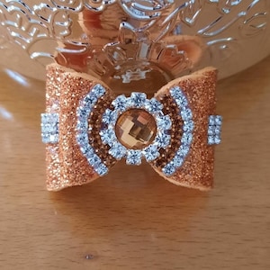 Puede incluir: Un lazo naranja brillante con un gran strass transparente en el centro. El lazo está decorado con strass más pequeños alrededor de los bordes.