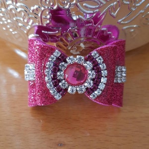 Puede incluir: Un lazo rosa brillante con un grupo central de pedrería transparente y rosa. El lazo presenta una textura brillante y está adornado con adornos de pedrería adicionales en cada lado, creando un accesorio glamuroso.
