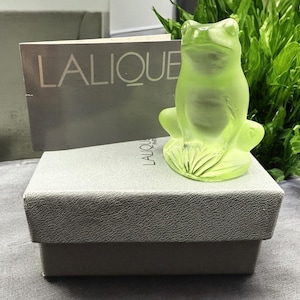 Puede incluir: Una figurita de rana de vidrio verde de Lalique, sentada en una caja gris con el logotipo de Lalique.