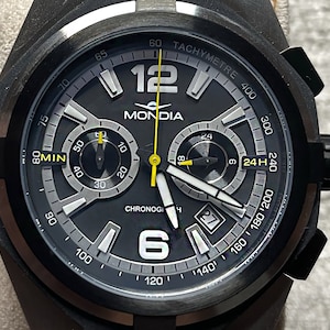Può includere: Un orologio da polso cronografo nero con quadrante argentato e dettagli gialli. Il quadrante dell'orologio presenta il nome del marchio "MONDIA" e le parole "TACHYMETRE" e "CHRONOGI".