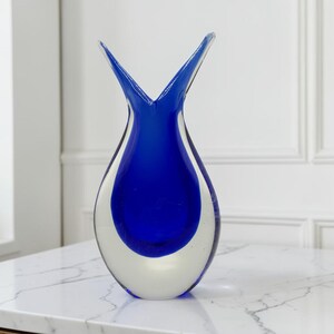 May include: A teardrop-shaped glass vase featuring a vibrant cobalt blue center encased in clear glass. The vase has a distinct V-shaped opening at the top, creating two upward-pointing points. The design is smooth, polished, and transparent.