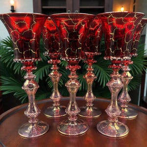 Könnte beinhalten: Sechs rote und goldfarbene Weingläser. Die Gläser haben ein ausgestelltes Schalen-Design mit einem goldenen Muster, einen dekorativen Stiel und einen runden Fuß. Sie stehen auf einem runden Holztisch.