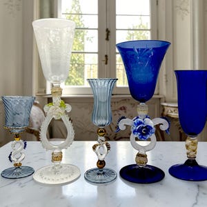 May include: An assortment of ornate, colorful glass goblets. The collection includes clear, light blue, and deep blue glasses with decorative bases and stems. Some feature floral and swirl designs.