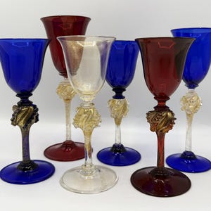 Puede incluir: Un conjunto de seis copas de cristal decorativas con cuencos acampanados en azul intenso, rojo oscuro y cristal transparente. Cada tallo presenta un adorno de cristal texturizado con motas doradas sobre una base circular, formando un juego.