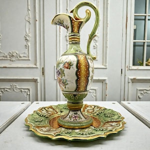 Può includere: Un'antica brocca in ceramica ornata con un vassoio decorativo abbinato. La brocca presenta un manico e un beccuccio curvi, con dettagli floreali e dorati su sfondo crema. Il vassoio è verde e oro, con un bordo smerlato.