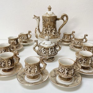 Puede incluir: Un juego de té de porcelana vintage en color crema y dorado. El juego incluye una tetera, un azucarero, una jarra de crema y 10 tazas y platillos. Cada pieza presenta un delicado diseño floral con querubines y un borde dorado.