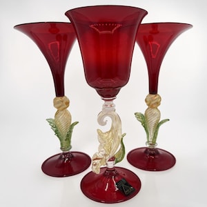 Può includere: Tre calici in vetro rosso rubino con bordi svasati e steli trasparenti attorcigliati, caratterizzati da decorazioni a forma di foglie in vetro dorato e verde. Ogni calice ha una base circolare e una piccola etichetta rettangolare nera sul davanti.