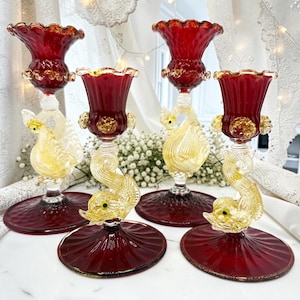 Puede incluir: Cuatro candelabros de cristal ornamentados. Cada uno presenta una copa de color rojo rubí con borde ondulado, un tallo transparente con diseño de delfín dorado y amarillo, y una base roja rubí. Decorativo.