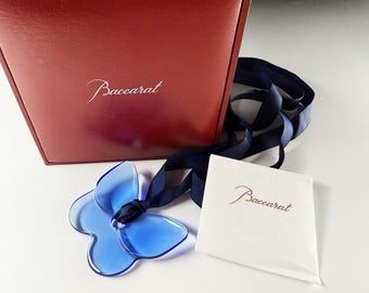 May include: A blue crystal butterfly pendant on a dark blue ribbon, accompanied by a red Baccarat box and a small white card with the Baccarat logo printed in red. The pendant features a smooth, translucent finish and a central hole for the ribbon.