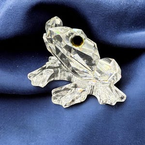 May include: A crystal frog figurine with a clear, faceted body and a single black eye. The frog is sitting on a blue fabric surface.