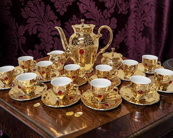 May include: A gold-colored tea set with a teapot, creamer, sugar bowl, and multiple teacups and saucers. Each piece is adorned with red accents. The set is arranged on a wooden surface.