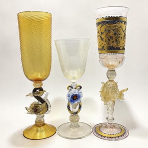 Puede incluir: Tres copas de vino decorativas. La copa de la izquierda es ámbar con un diseño en espiral y una base en forma de cisne. La copa del medio es transparente con una base floral. La copa de la derecha tiene un diseño dorado y azul.