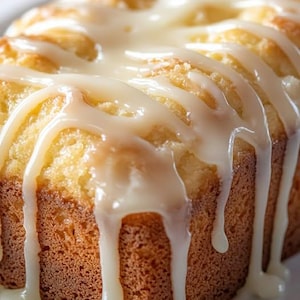 May include: A close-up of a loaf of golden-brown cake with a white glaze drizzled over the top. The glaze is thick and creamy, and it is dripping down the sides of the cake. The cake appears to be freshly baked and moist.