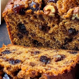 May include: A close-up of a loaf of fruitcake with a slice cut off. The cake is a golden brown color and is studded with dark raisins and walnuts. The texture is dense and moist, with a slightly rough surface.