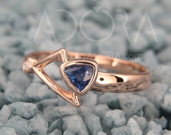 Puede incluir: Un anillo de oro rosa con una piedra preciosa azul triangular y un pequeño acento de diamante. El anillo está colocado sobre un fondo de pequeñas piedras azules.