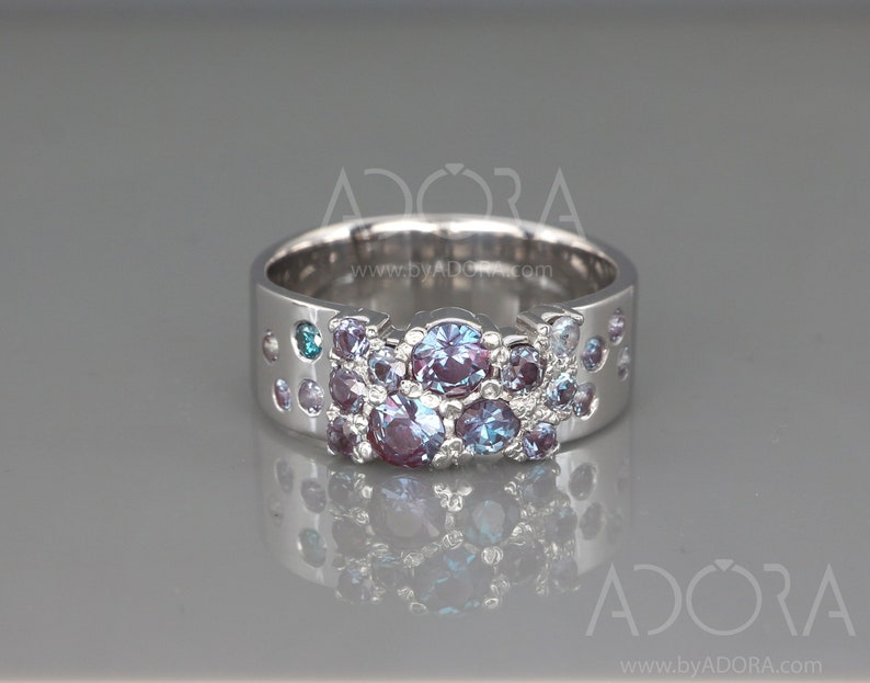 May include: A silver ring with a wide band, adorned with a cluster of round gemstones in shades of purple and blue, with teal highlights. The ring has a contemporary design, incorporating small, decorative perforations.