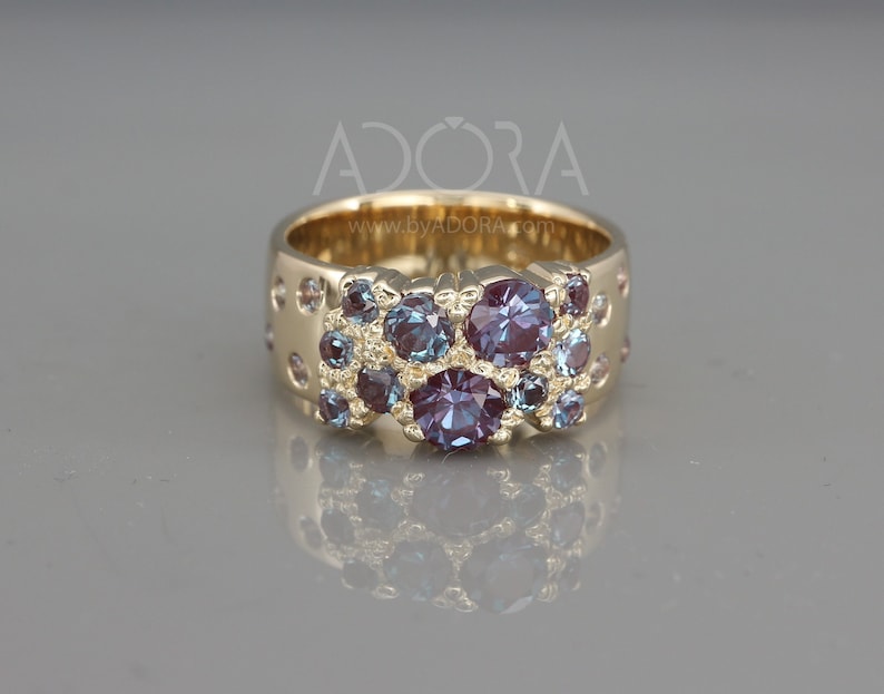 May include: A gold band ring with a cluster of purple and blue gemstones. The ring is set with a variety of sizes and shapes of stones, creating a unique and eye-catching design. The ring is made of 14k gold and is available in a variety of sizes.