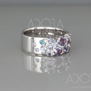 May include: A silver ring with a wide band, adorned with a cluster of round gemstones in shades of purple, blue, and teal. The ring's design includes smaller, scattered stones along the band, creating a sparkling effect. The ring is displayed on a reflective surface.