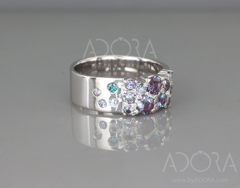 May include: A silver ring with a wide band, adorned with a cluster of round gemstones in shades of purple, blue, and teal. The ring's design includes smaller, scattered stones along the band, creating a sparkling effect. The ring is displayed on a reflective surface.