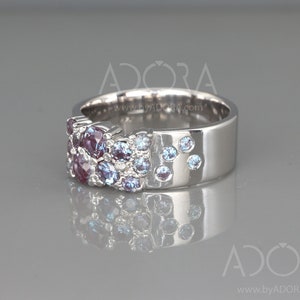 May include: A silver ring featuring a cluster of purple and blue gemstones. The ring has a wide band with a smooth, polished finish. The gemstones are arranged in a textured pattern on the top of the ring, with some scattered on the side.