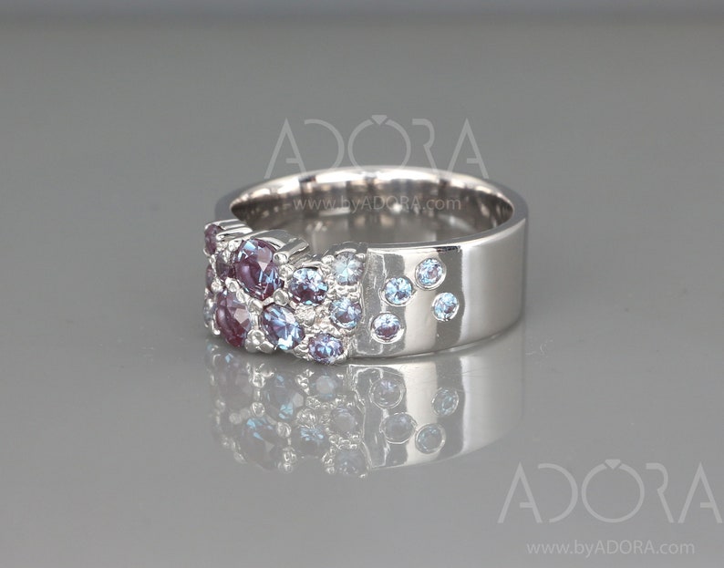 May include: A silver ring featuring a cluster of purple and blue gemstones. The ring has a wide band with a smooth, polished finish. The gemstones are arranged in a textured pattern on the top of the ring, with some scattered on the side.