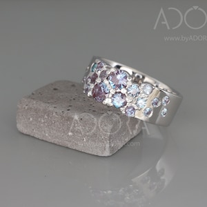 May include: A silver ring featuring a cluster of round gemstones in shades of purple and blue. The ring is presented on a small, grey, rectangular stone. The ring's design is modern and elegant. The gemstones are set across the ring's surface.