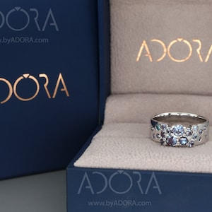 May include: A silver ring featuring a collection of blue and purple gemstones. The ring is presented in a dark blue box, branded with "ADORA" in gold. The ring's design includes small, circular cutouts around the band.