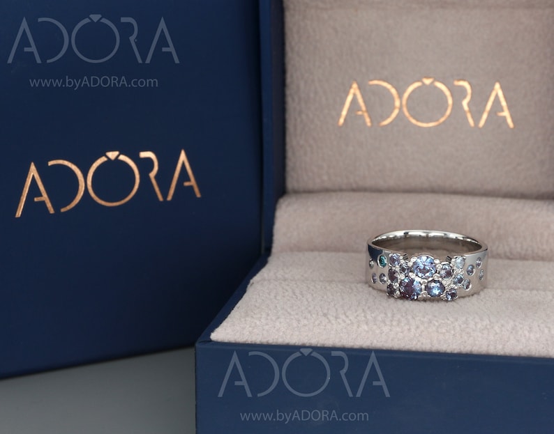 May include: A silver ring featuring a collection of blue and purple gemstones. The ring is presented in a dark blue box, branded with "ADORA" in gold. The ring's design includes small, circular cutouts around the band.